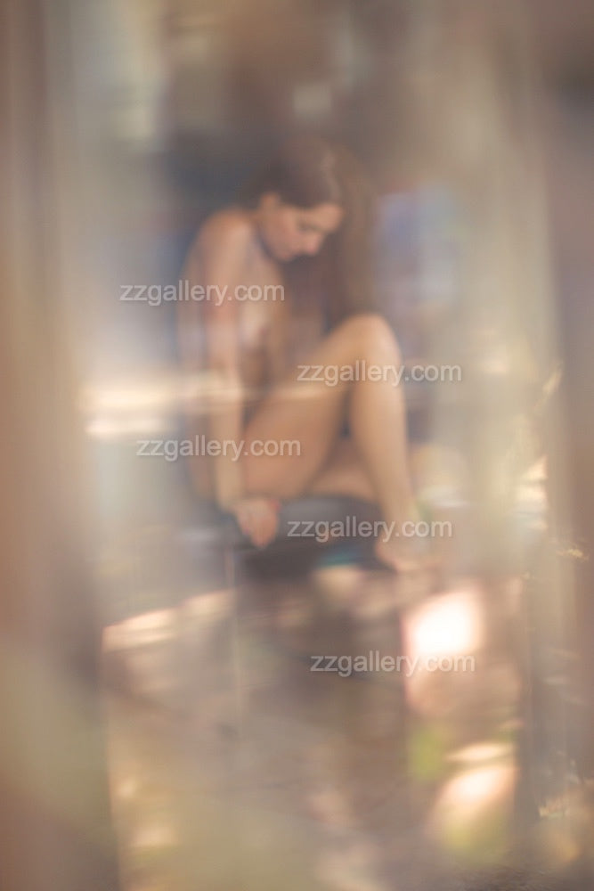 Blurry fine art photograph of a golden-painted figure seated in soft natural light, viewed through an ethereal veil of glass and bokeh.