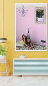 Whimsical fine art photograph of a woman in a yellow dress sitting beside a vacuum cleaner filled with candy, set against a pink wall – surreal wall art for playful interiors and modern home décor.