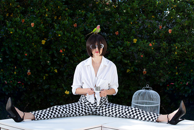 Surreal pop art fine art photograph by Zack Zublena titled Coffee Time, featuring a woman in checkered pants sipping coffee with a parrot on her head, heart-shaped sunglasses, and an empty birdcage in the background.